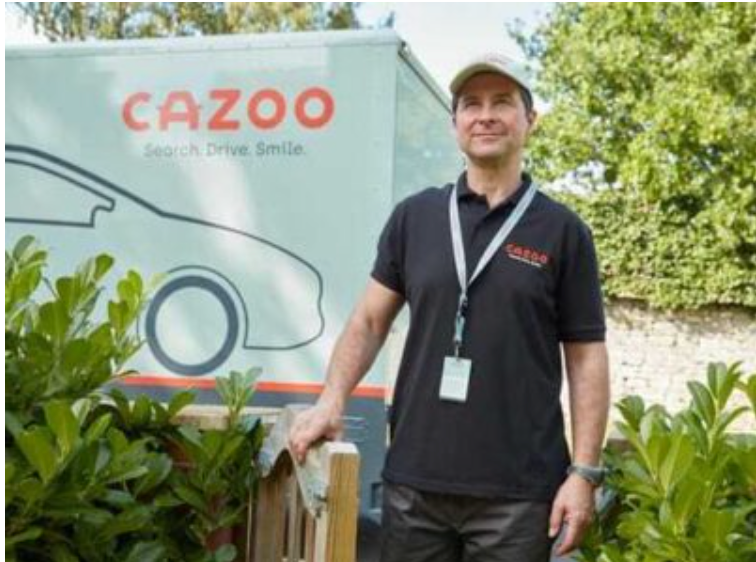 Man in black polo shirt standing beside Cazoo delivery van with greenery background