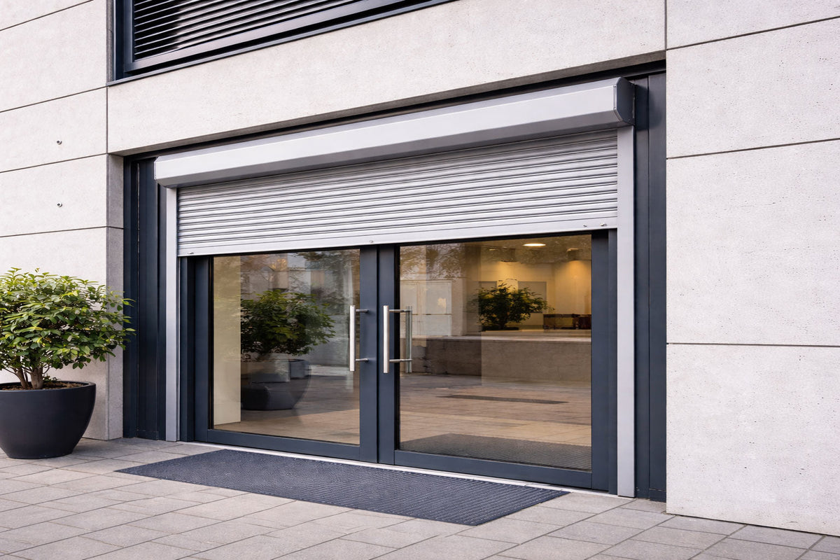 Silver aluminium roller shutter partially open over glass double doors of a modern business entrance