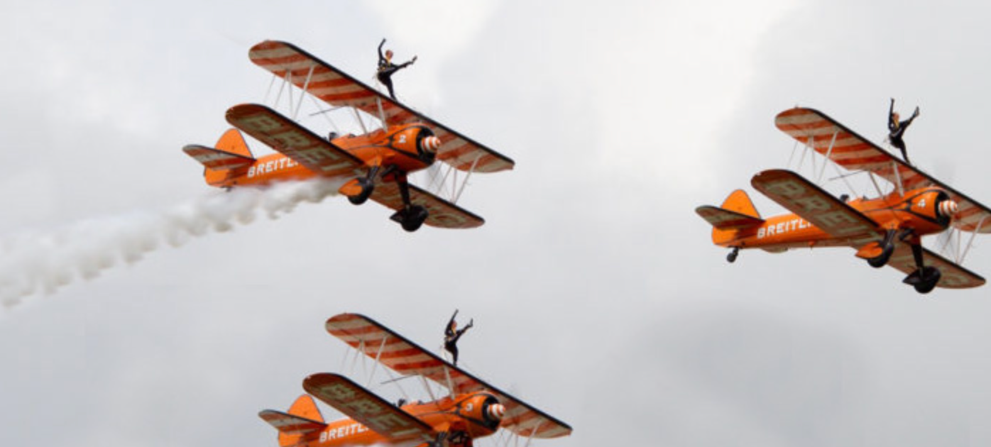 Orange biplanes with wing walkers performing aerial stunts in cloudy sky