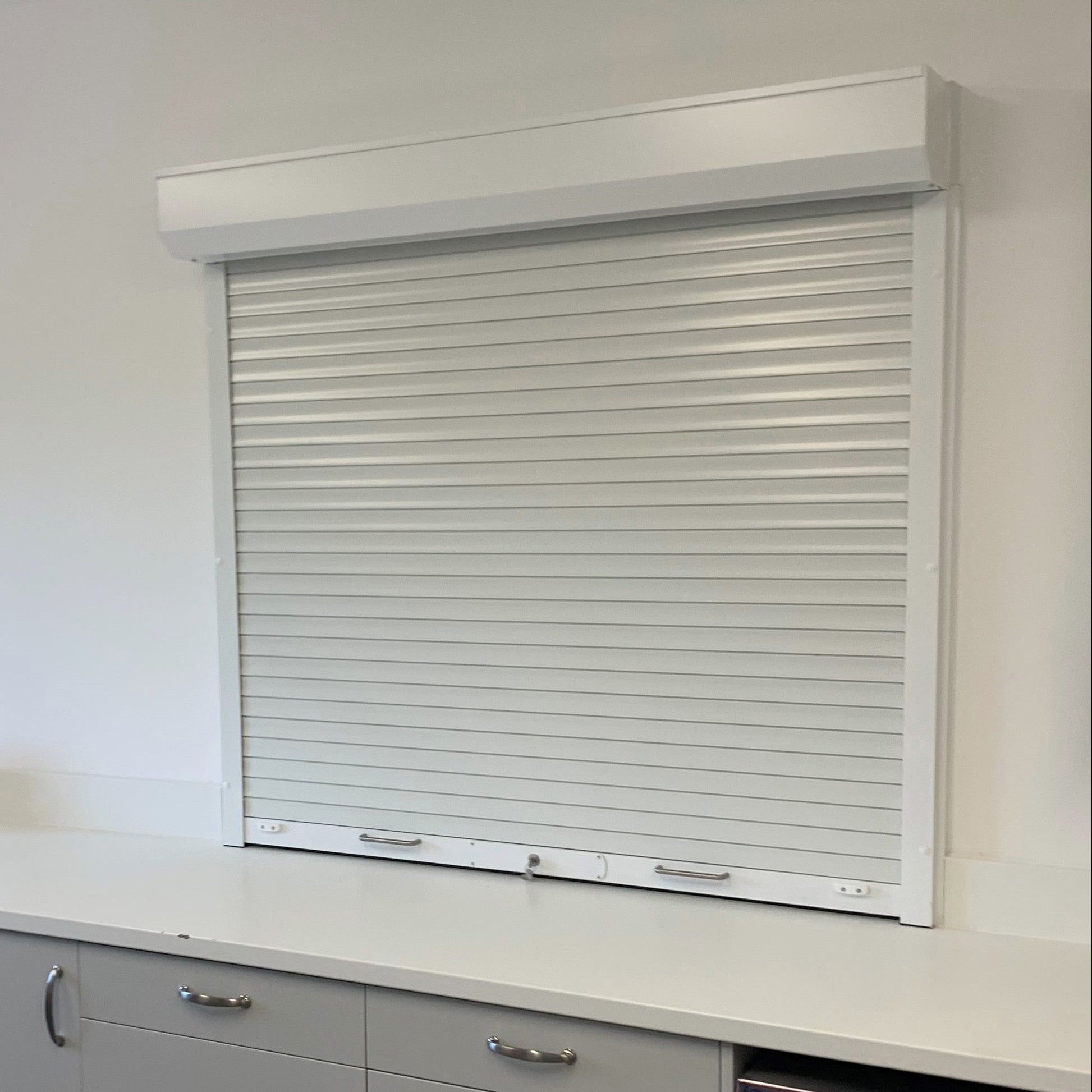 A white roller shutter window blind installed on a wall above a modern kitchenette counter with light gray cabinets and a stainless steel dishwasher.