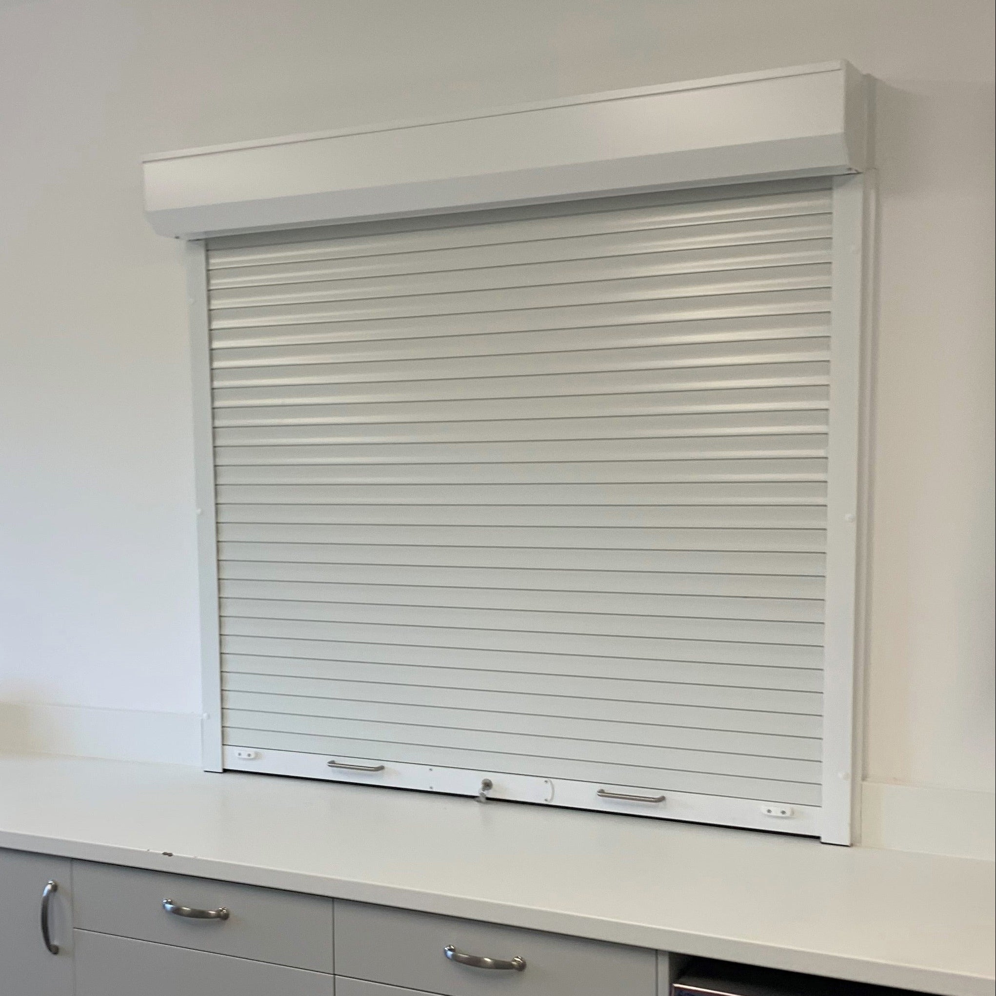 A white roller shutter window blind installed on a wall above a modern kitchenette counter with light gray cabinets and a stainless steel dishwasher.